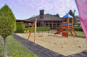 A playground with swings and a slide, a wooden playhouse, and a building with a chimney in the background.