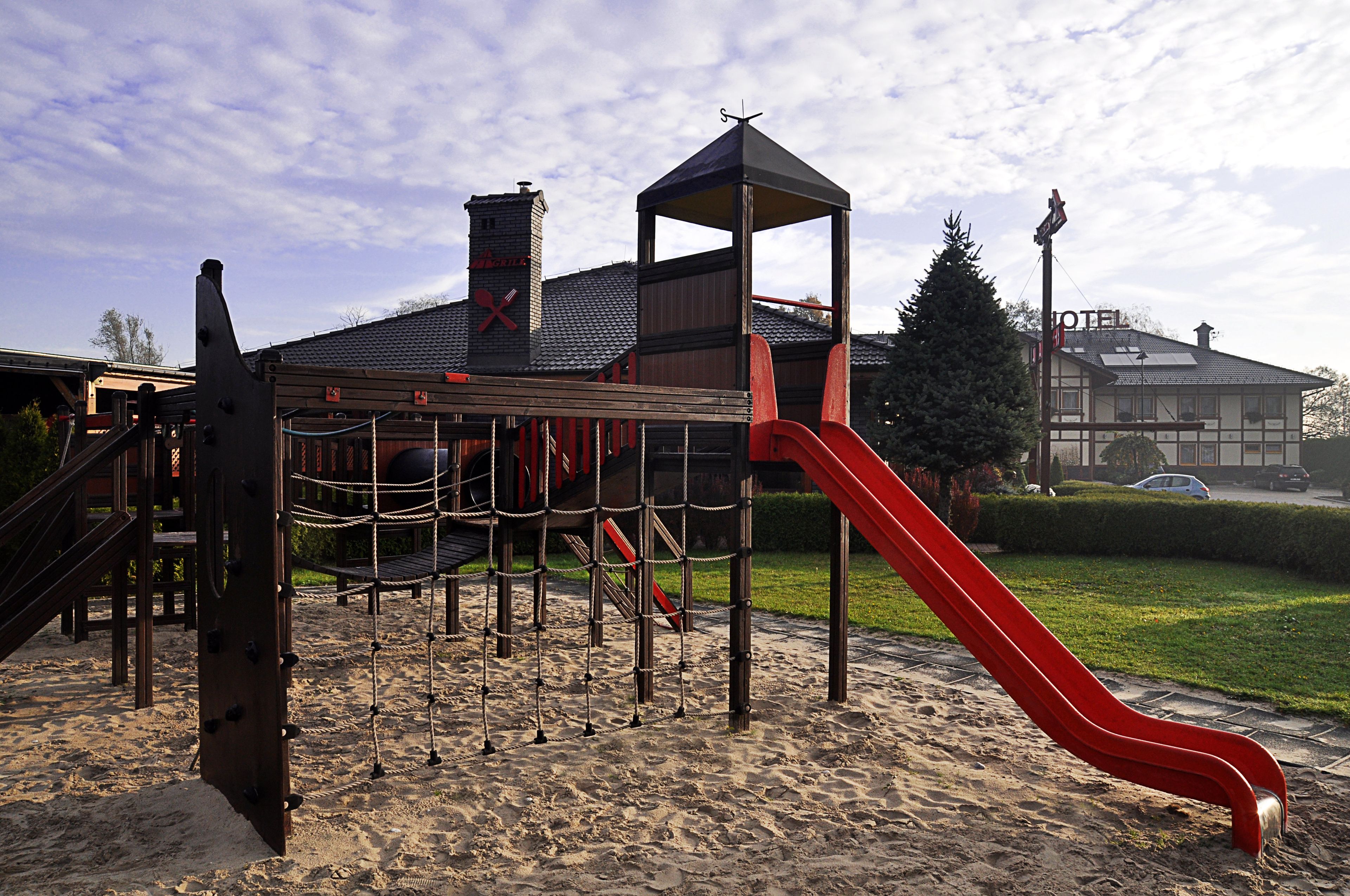 Ein Spielplatz mit einer roten Rutsche und Klettergerüsten, vor einem Gebäude mit Schornstein und einem Schild mit der Aufschrift 'HOTEL'.