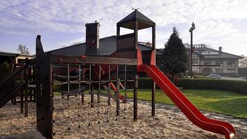 Ein Spielplatz mit einer roten Rutsche und Klettergerüsten, vor einem Gebäude mit Schornstein und einem Schild mit der Aufschrift 'HOTEL'.