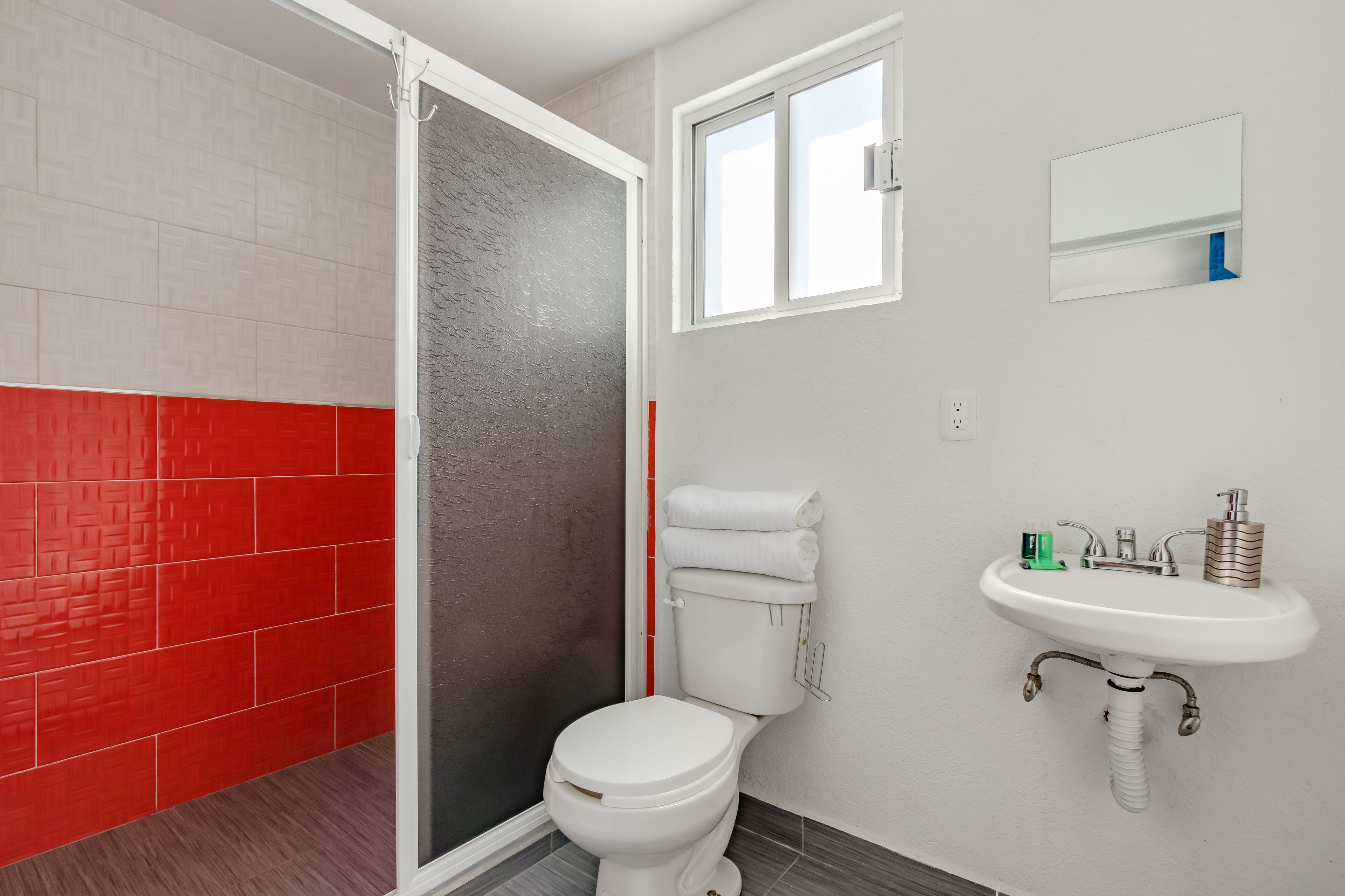Een badkamer met een wit toilet, een wastafel met spiegel erboven en een douchegedeelte met een betegelde muur in rood en grijs.