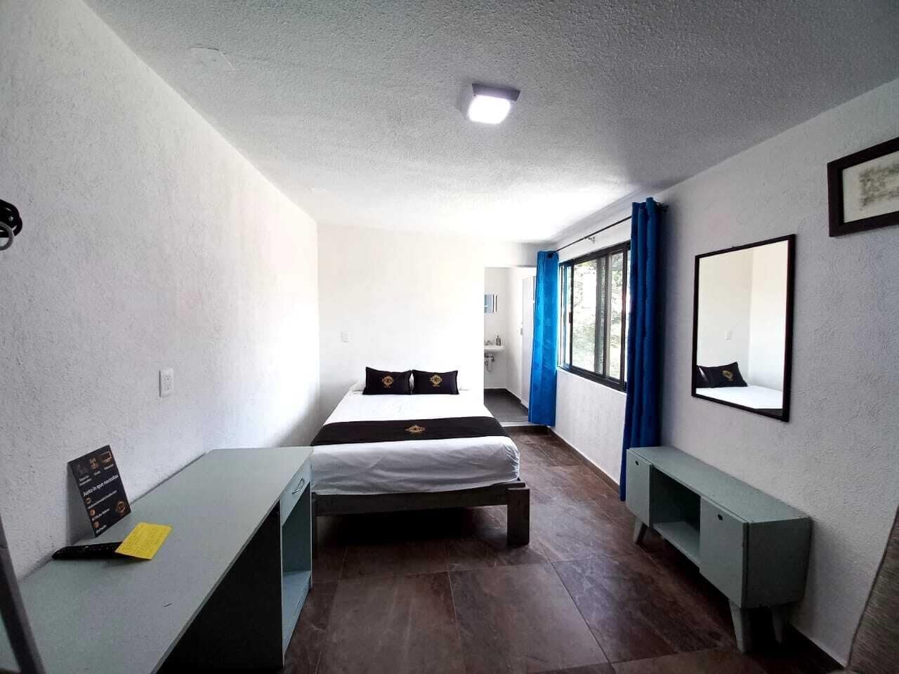 A compact hotel room with a bed, a desk, a bench, and a large window with blue curtains.