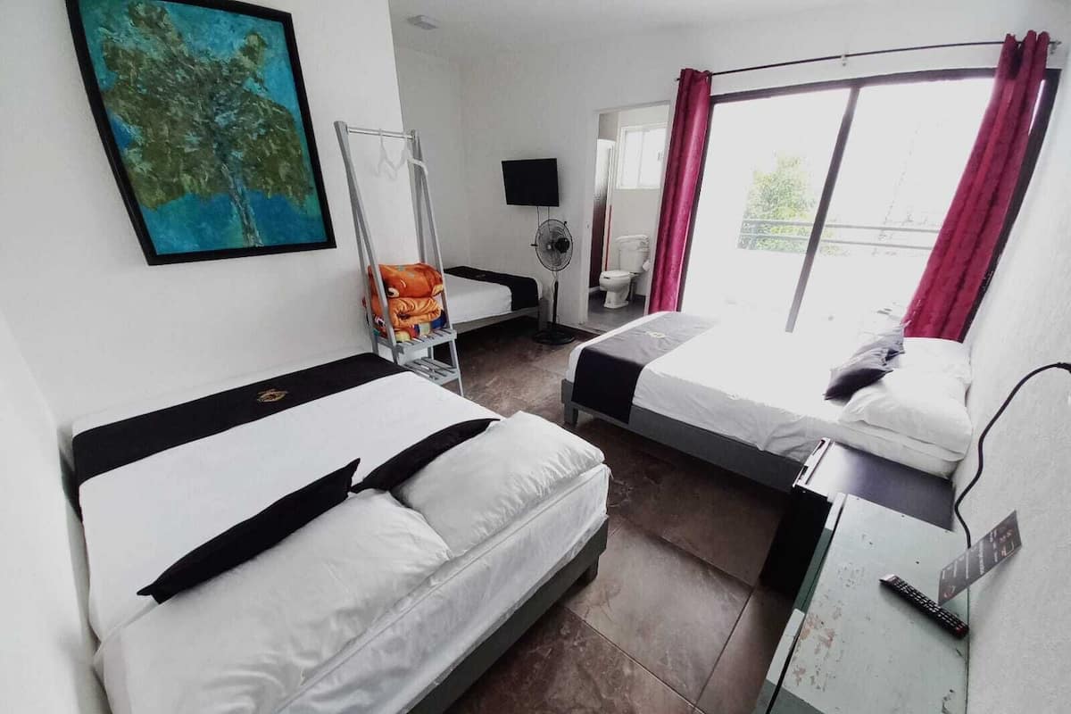 A hotel room with two beds, a TV, a fan, a window with curtains, and a painting on the wall.