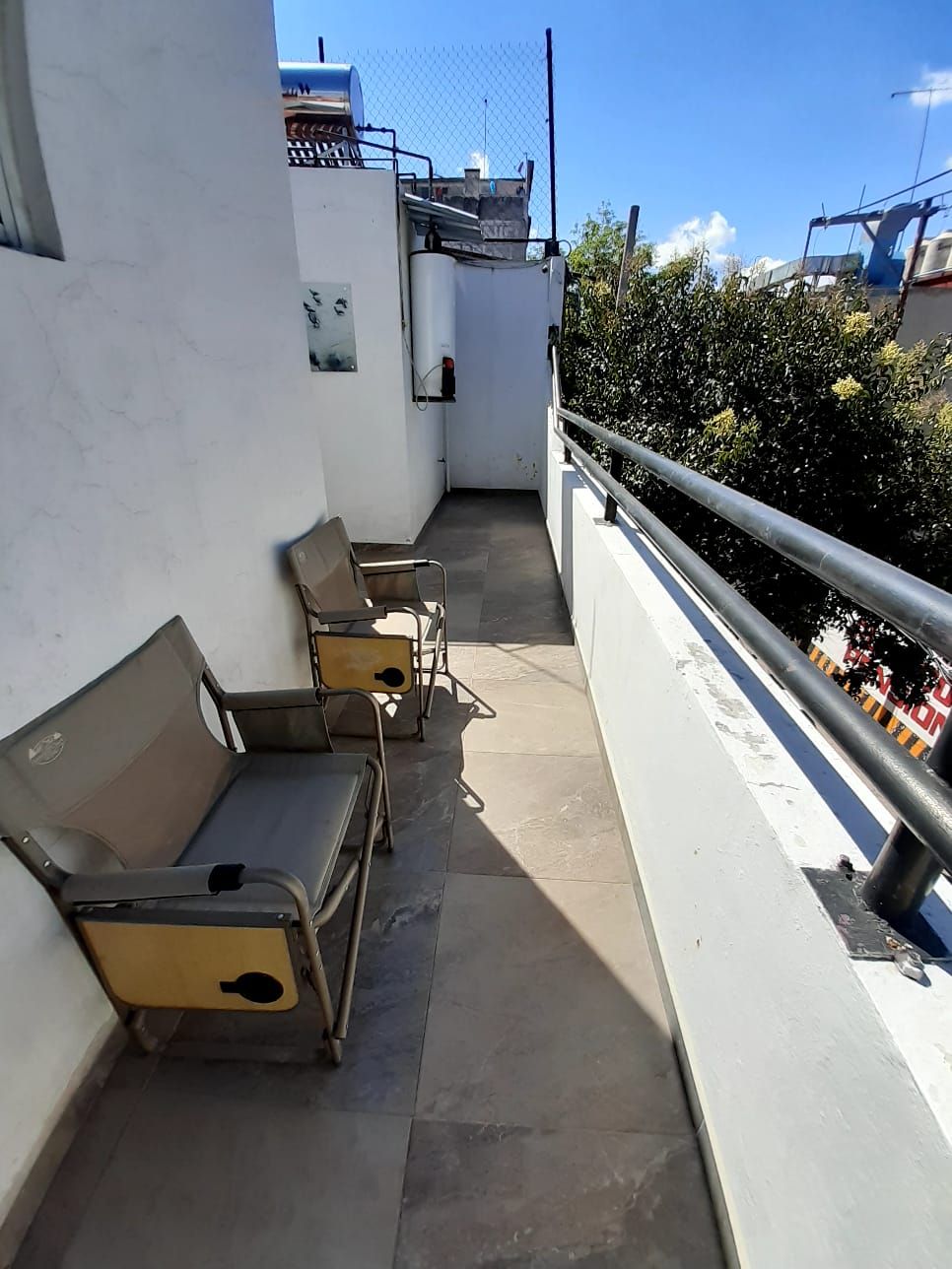 A balcony with two metal chairs and a bench, a metal railing, and a view of a building with a blue roof.