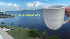 Une tasse blanche est tenue devant un paysage pittoresque comprenant une étendue d’eau, une zone herbeuse et une côte lointaine parsemée de bâtiments.