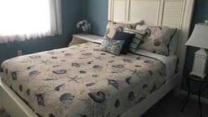 Un lit bien fait, avec une literie à motifs floraux, une tête de lit blanche et une table de chevet surmontée d’une lampe.