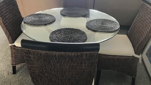 Une table ronde en verre, surmontée de sets de table en fibres tressées, entourée de chaises en osier dotées d’assises rembourrées et accompagnée d’un pouf assorti.