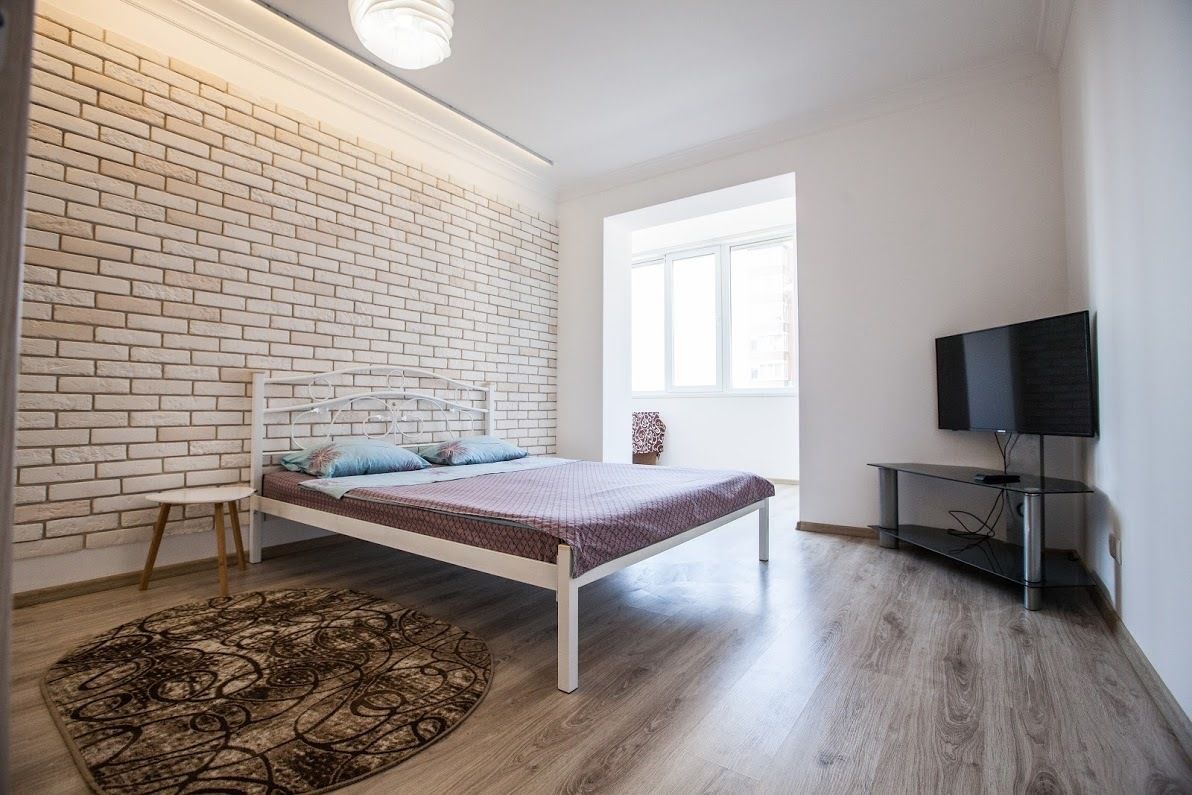 A bedroom with a white brick wall, a bed with a white frame, a TV on a stand, and a window providing natural light.