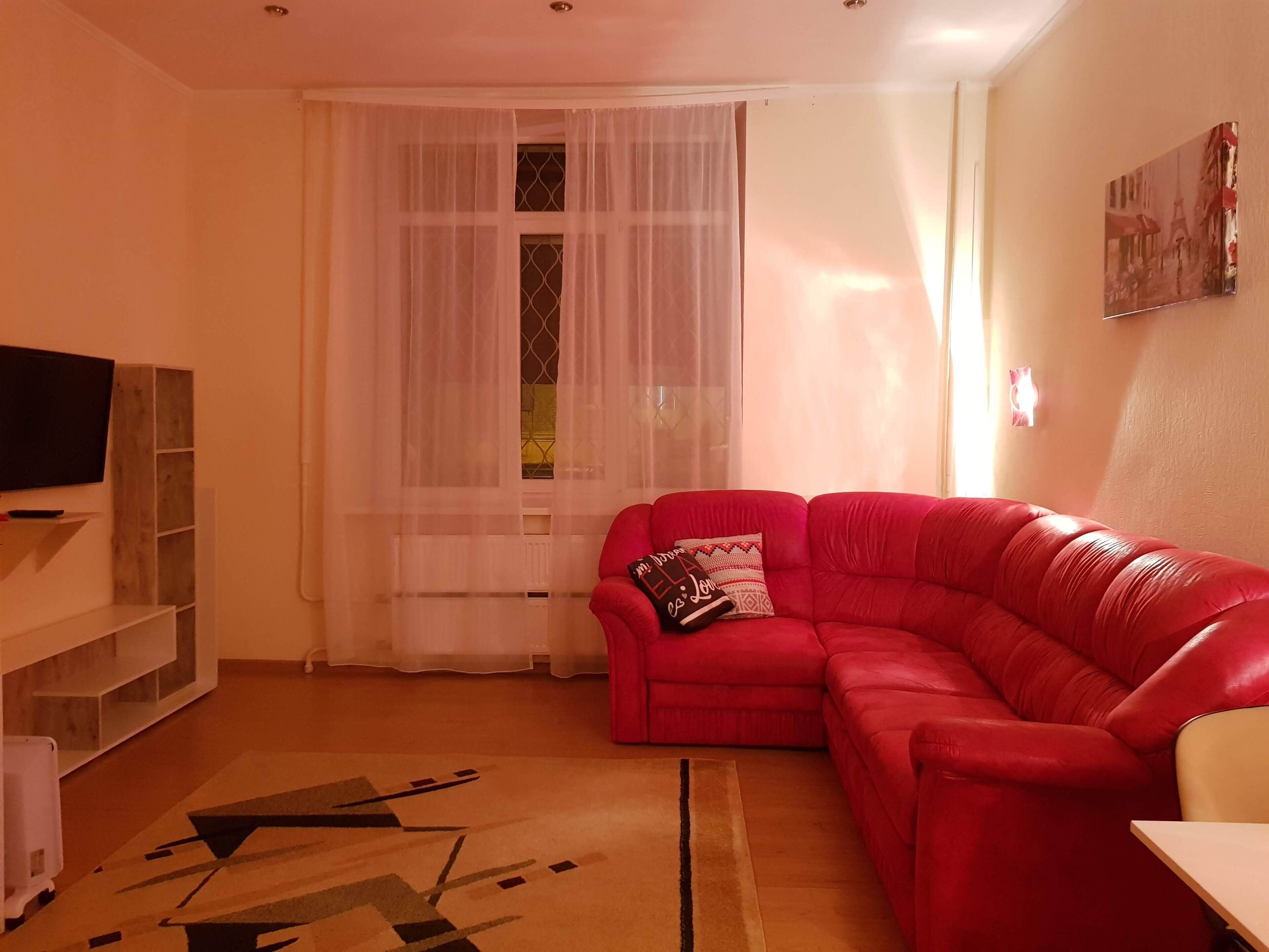 A living room with a red sofa, a television on a wooden stand, a window with sheer curtains, and a painting on the wall.