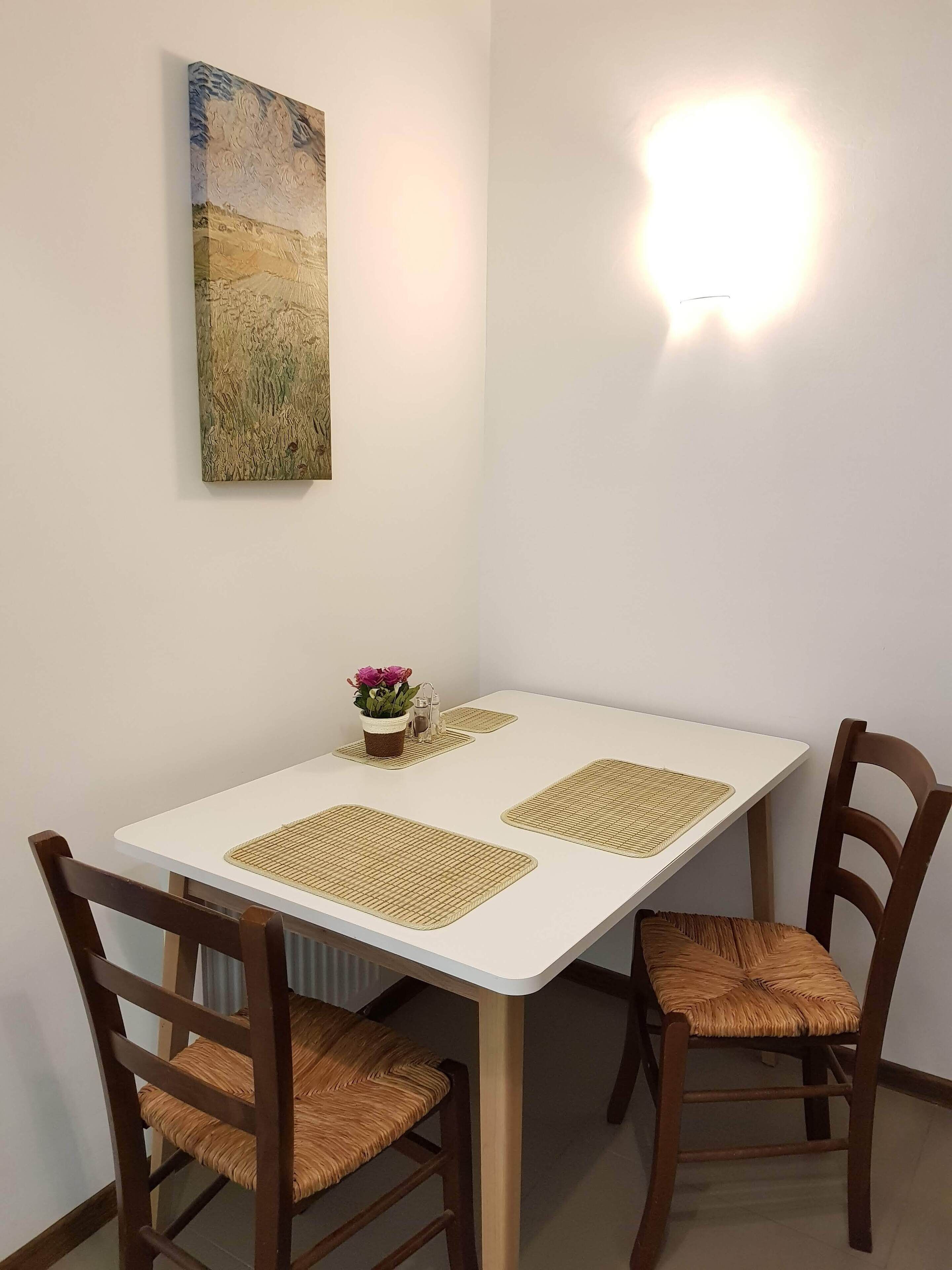 A dining table with four chairs, a painting on the wall, and a wall-mounted light fixture.