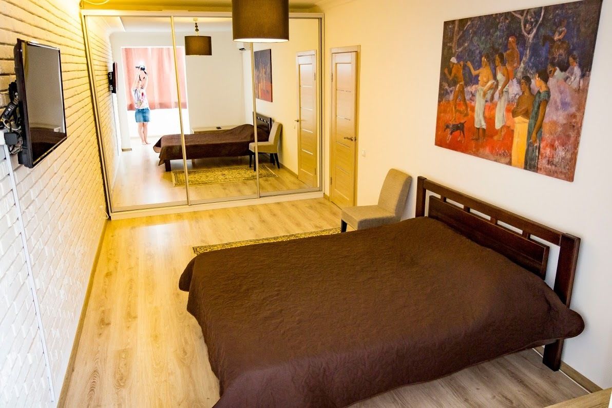 A bedroom with a wooden floor, a bed with a brown cover, a mirror, a painting, and a door leading to another room.