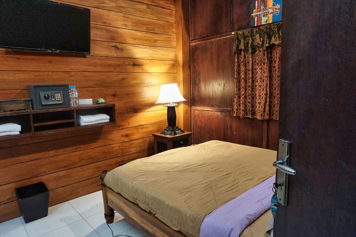 A wooden-paneled room with a bed, a TV mounted on the wall, a lamp, and a shelf with towels.