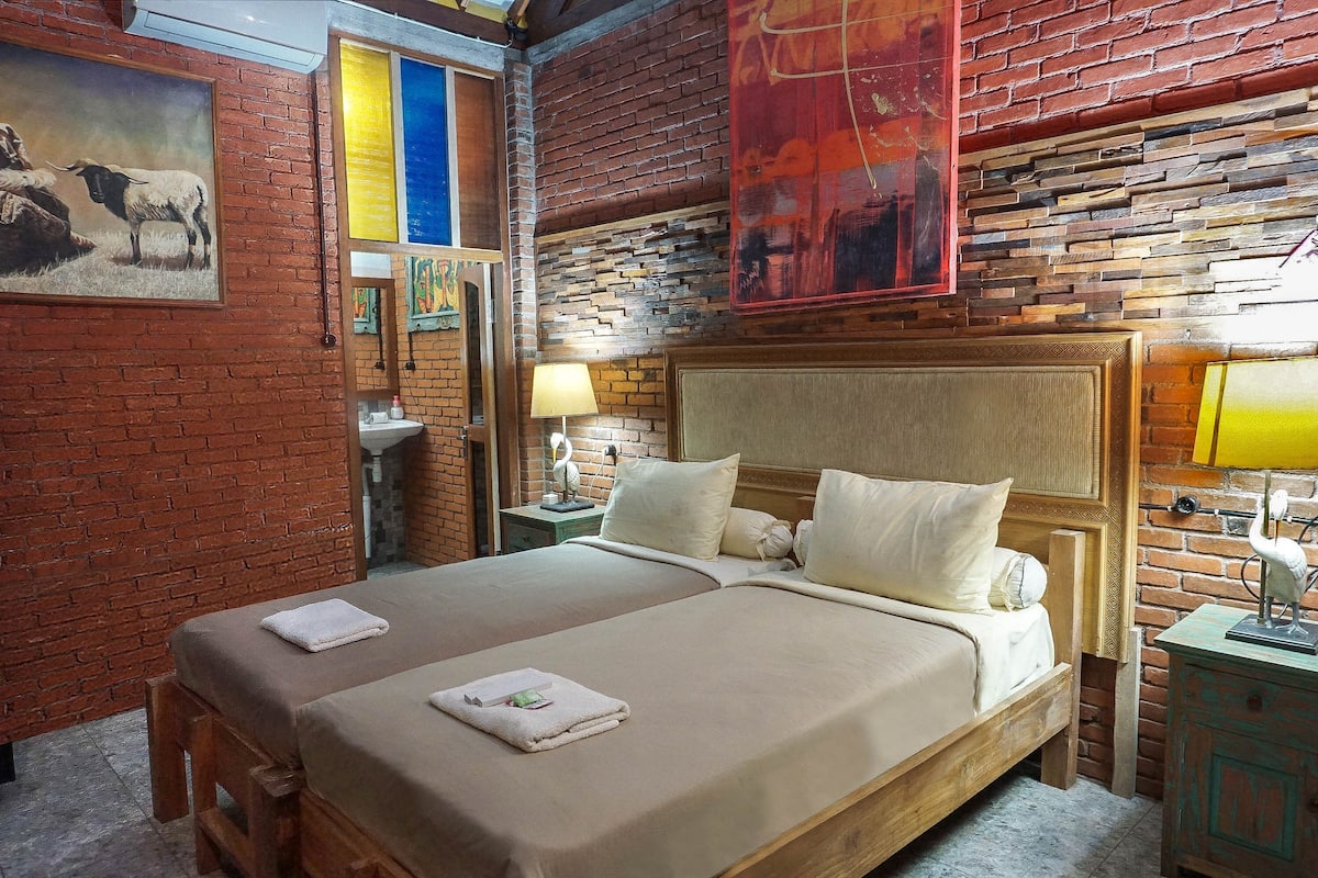 A hotel room with a brick wall, a large bed with white bedding, a wooden headboard, and a painting of a cow.