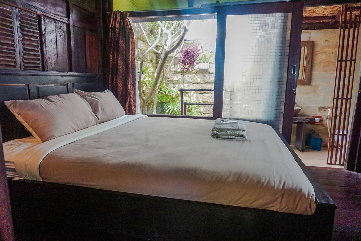 A neatly made bed with a white sheet and pillows, a wooden headboard, and a view of a garden through a glass door.