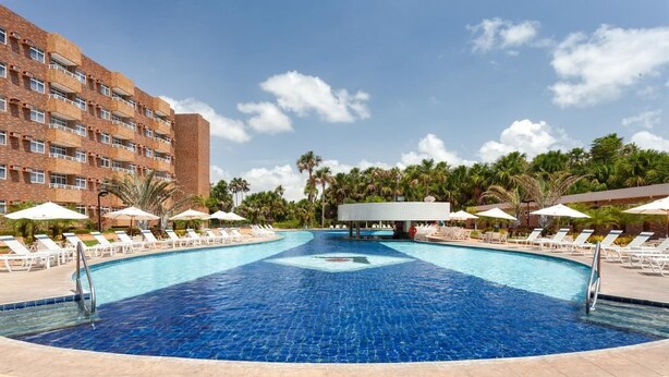 Una piscina con sillas de descanso y sombrillas, rodeada de palmeras y un edificio de hotel.