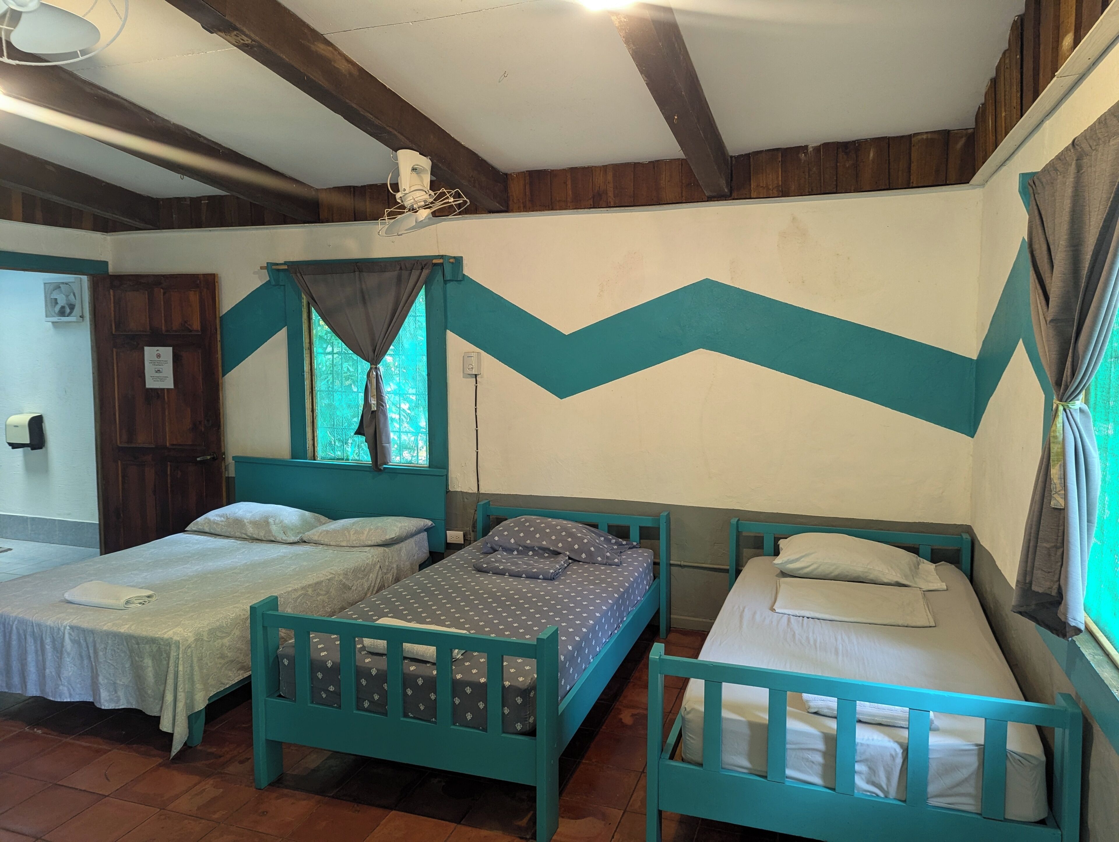A room with three wooden beds, a window with curtains, a ceiling fan, and a door with a sign.