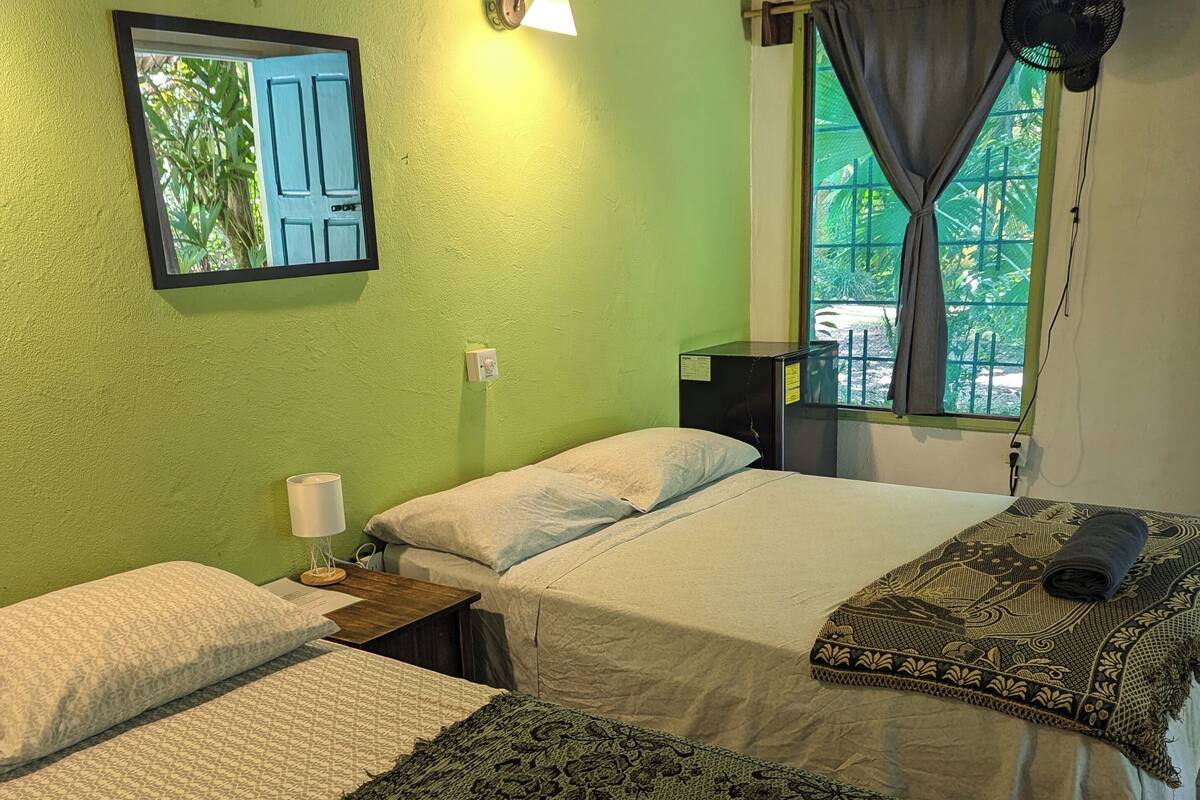 A room with two beds, a nightstand, a lamp, a mirror, a window with curtains, and a fan.