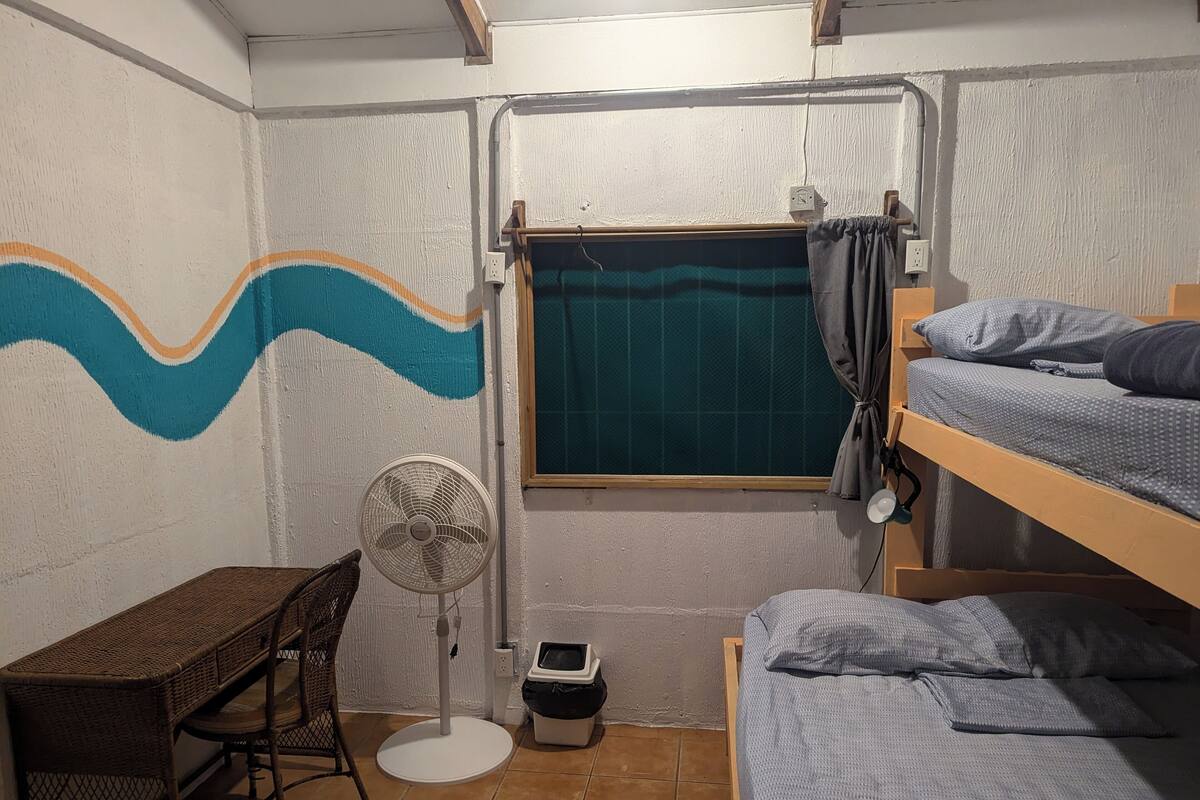 A dormitory room with bunk beds, a desk, a chair, a standing fan, a window with a curtain, and a wall with a wave pattern.