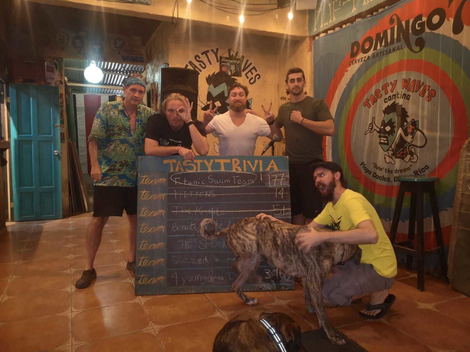Um grupo de pessoas posando em um bar, com um quadro-negro exibindo nomes de equipes e placares.