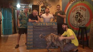 Um grupo de pessoas posando em um bar, com um quadro-negro exibindo nomes de equipes e placares.