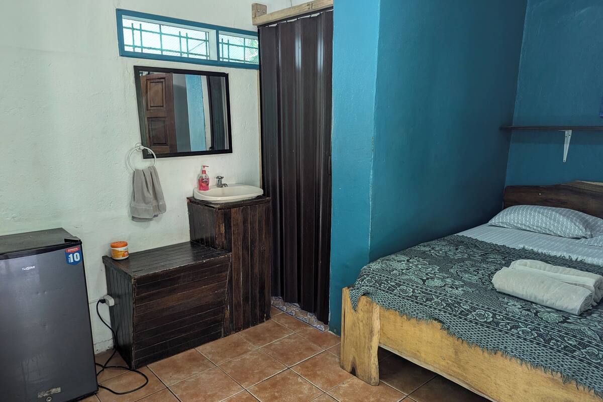A bedroom with a bed, a wooden dresser, a small sink, a mirror, and a refrigerator.