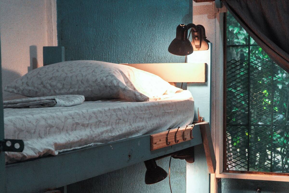 A bunk bed with a bedside lamp, a window with a curtain, and a view of greenery outside.