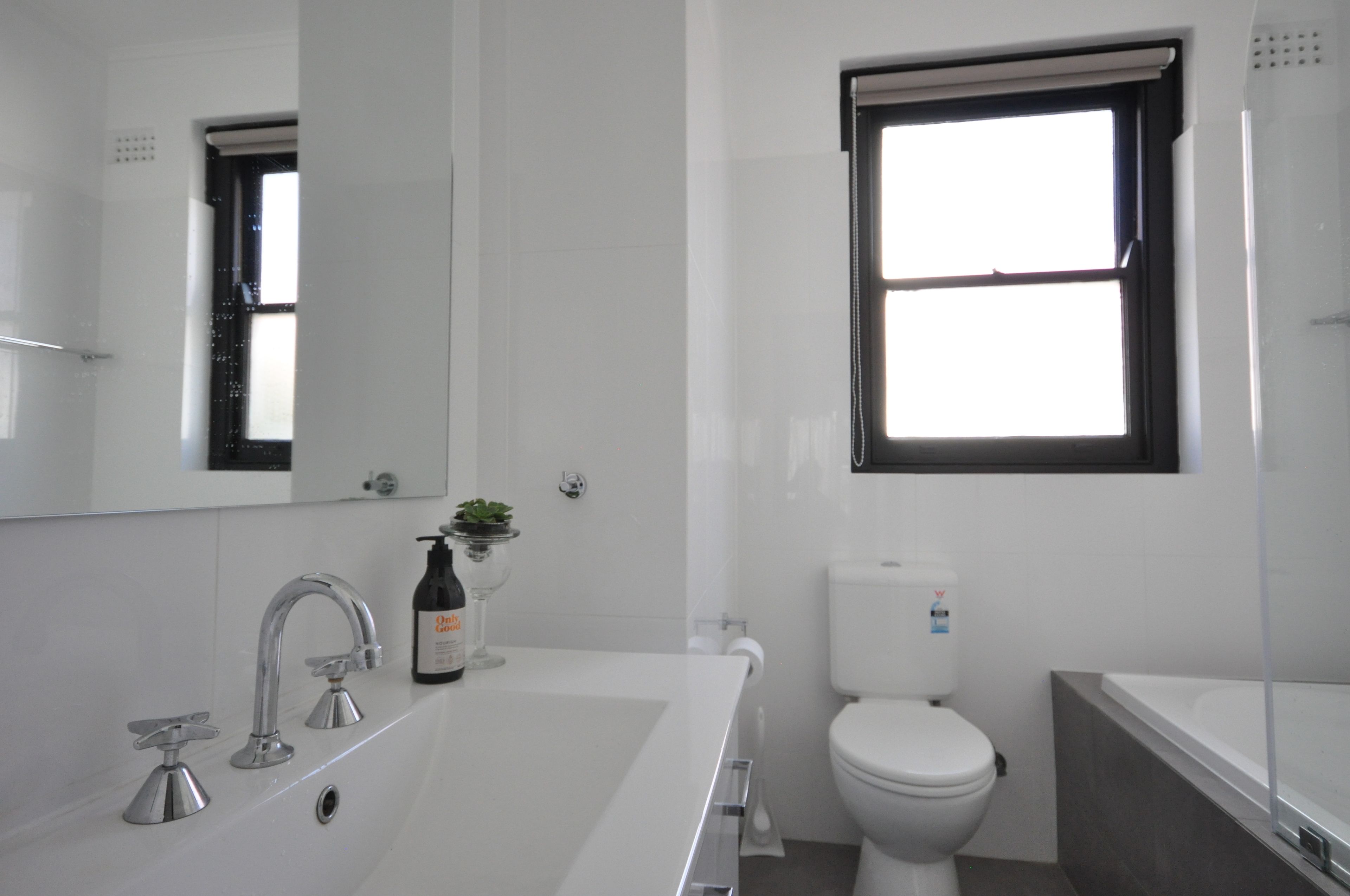 Een moderne badkamer met een witte wastafel, twee kraanwerk, een spiegel, een toilet en een raam.
