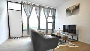 Una sala de estar moderna con un sofá gris, una mesa de centro de madera, un televisor de pantalla plana y un gran ventanal con cortinas.