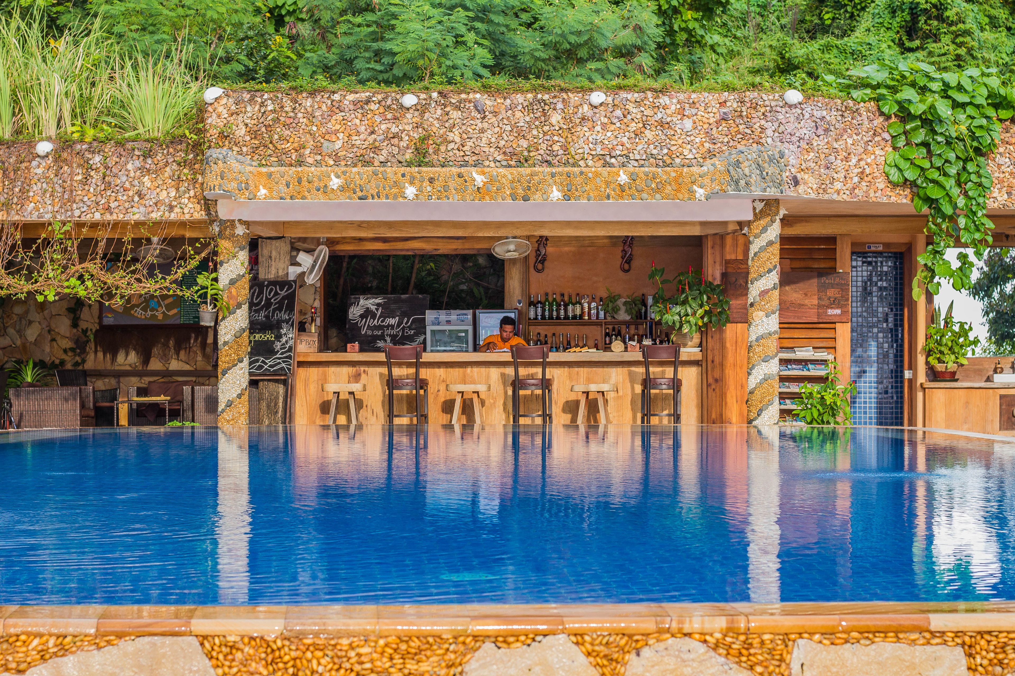 Un bar junto a la piscina con un fondo de pared de piedra, asientos de madera y un cantinero.