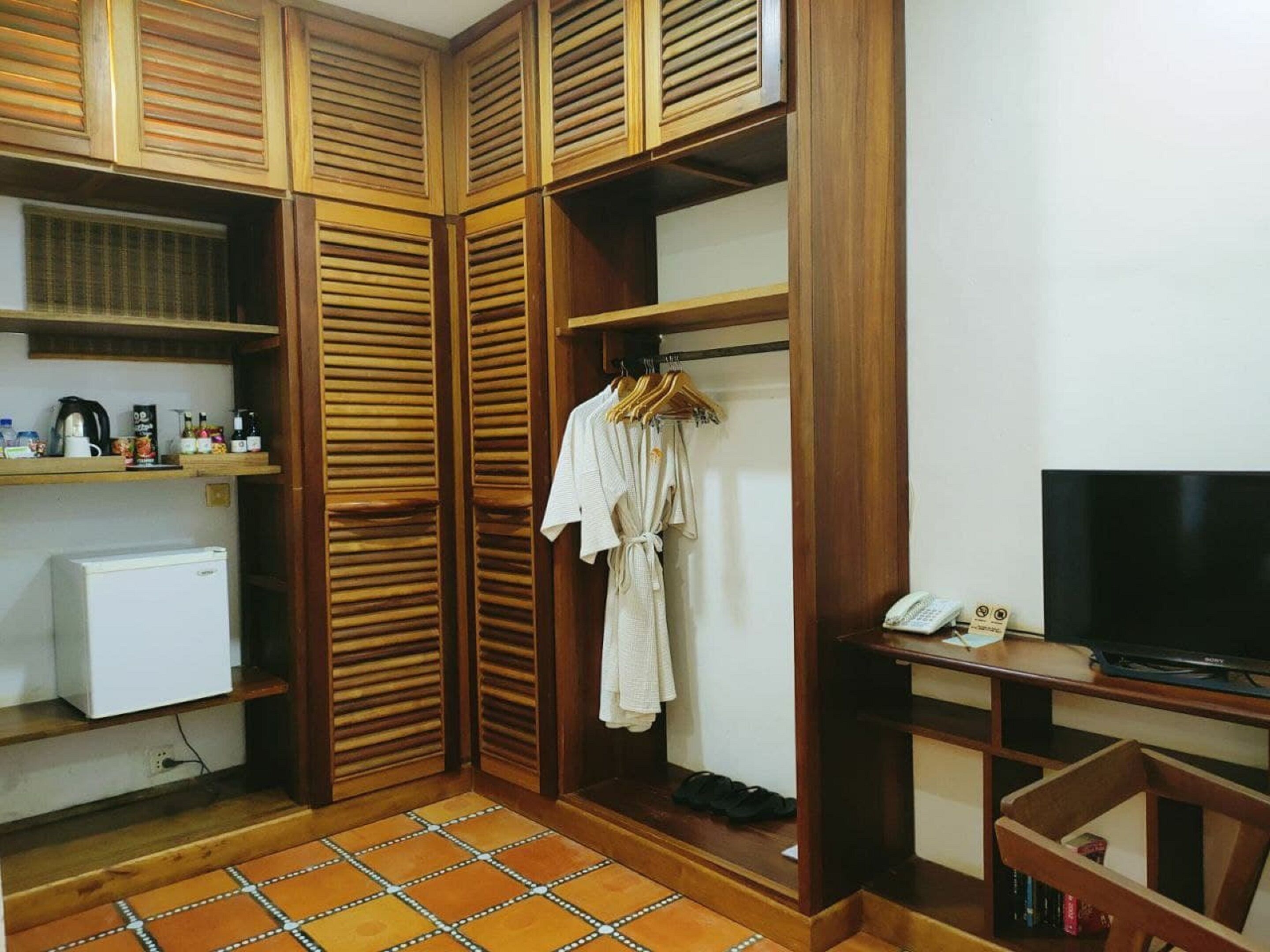 Una habitación compacta con un armario de madera, un refrigerador pequeño, un televisor y piso de baldosas.