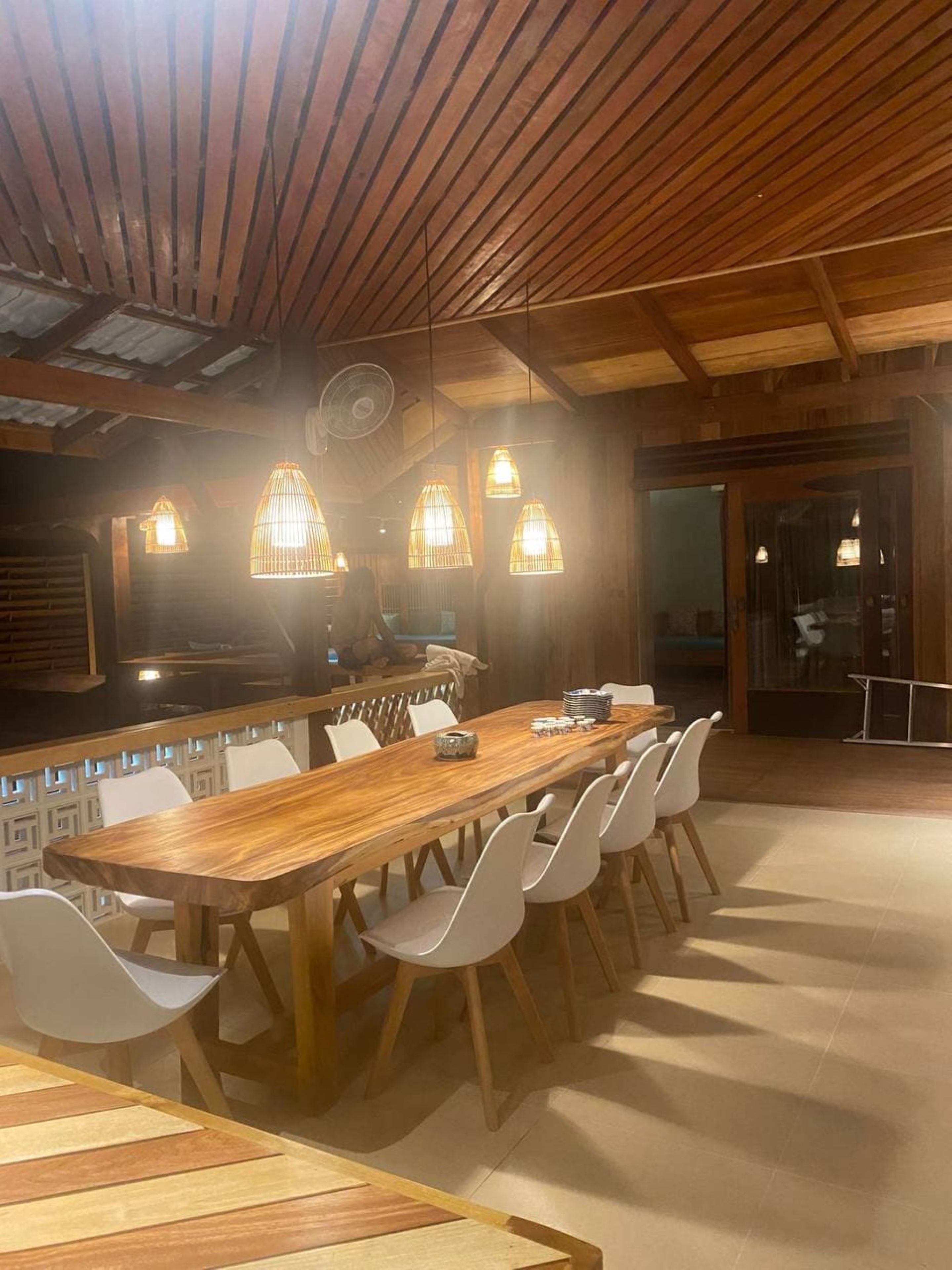 A wooden dining area with a long table and white chairs, a bar counter, and hanging pendant lights.