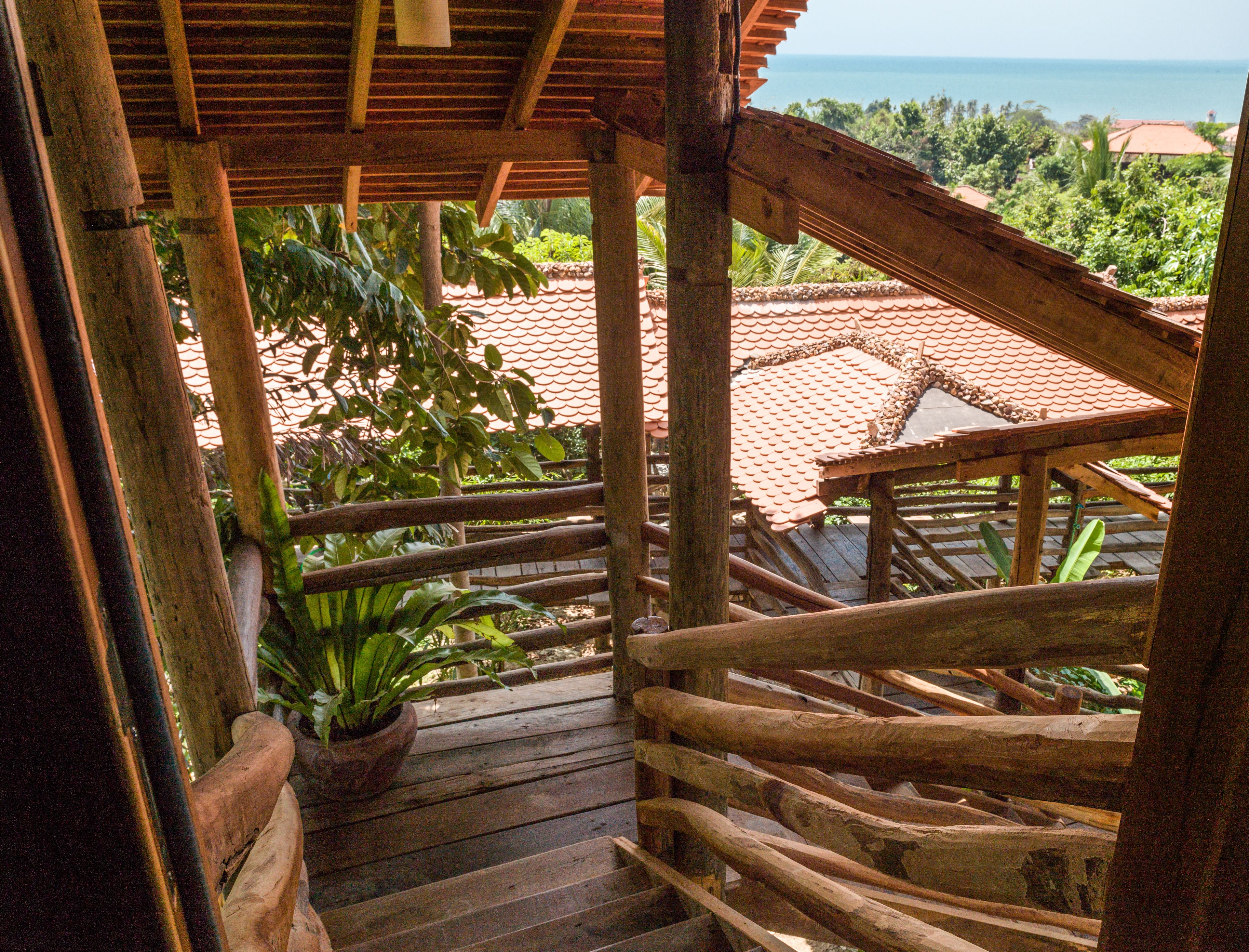 A wooden staircase leading to a covered outdoor area with a view of the sea and lush greenery.