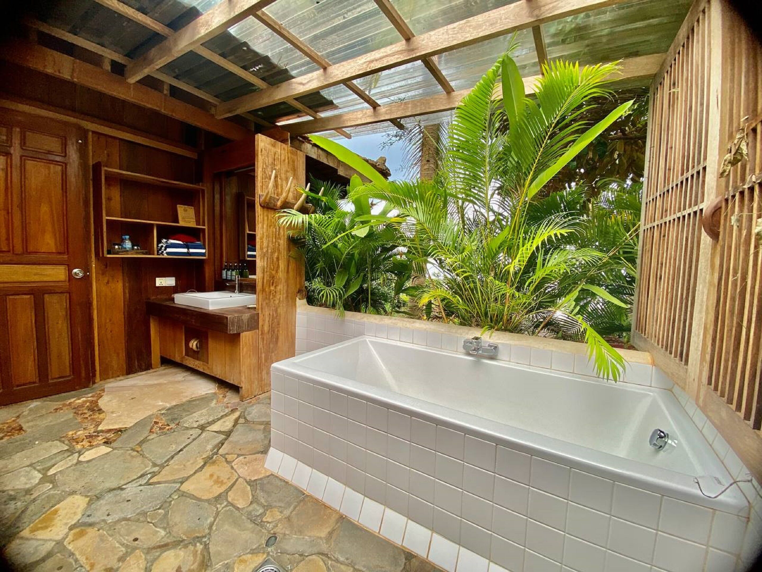 A bathroom with a stone floor, a bathtub, wooden cabinets, and a window with tropical plants.