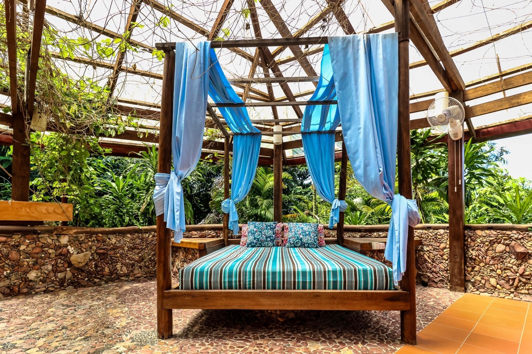 A wooden four-poster bed with blue drapes, a striped cushion, and a stone wall backdrop.