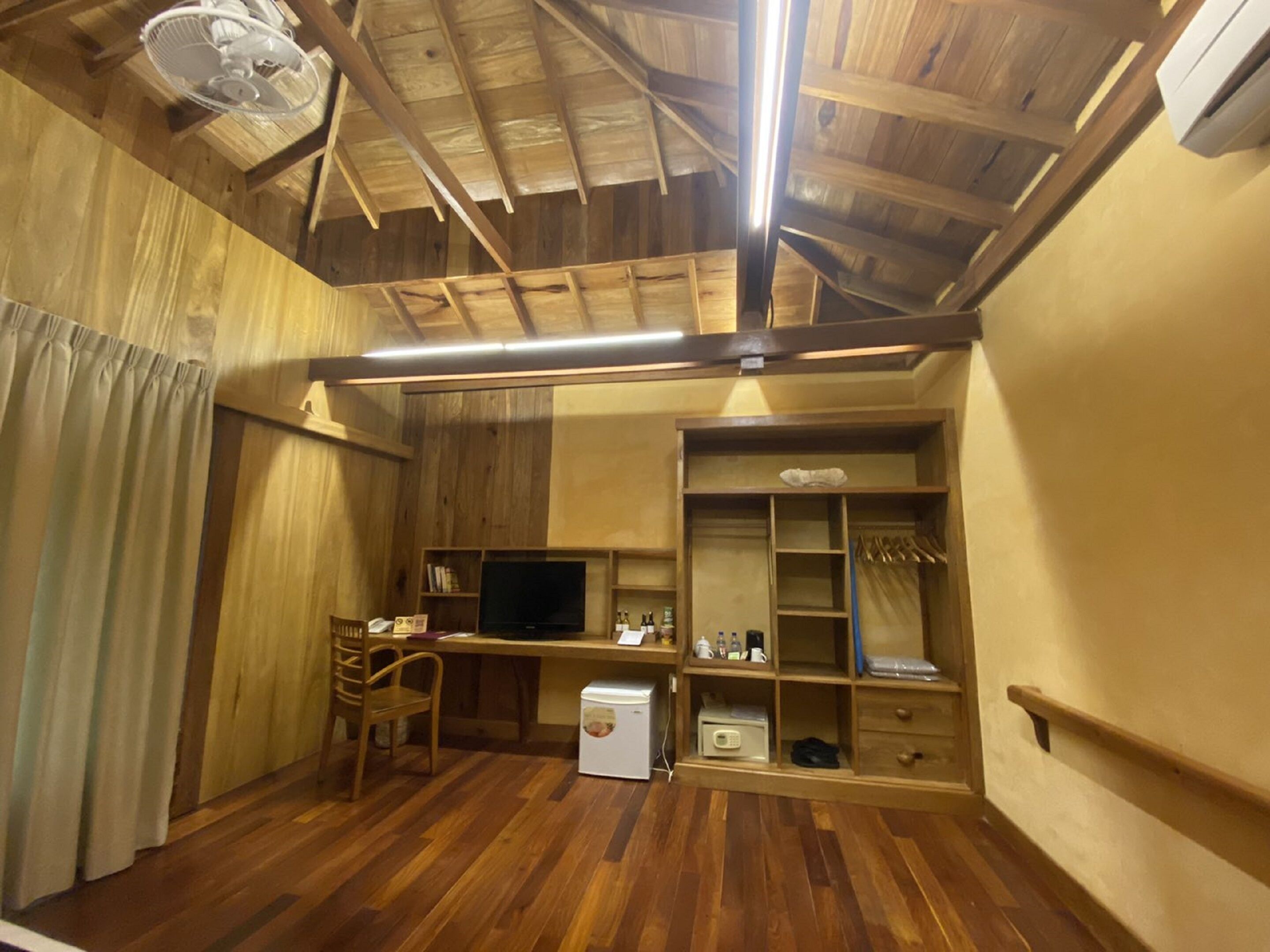 A compact room with wooden flooring, a ceiling fan, a television, a small refrigerator, and a wooden wardrobe with shelves.