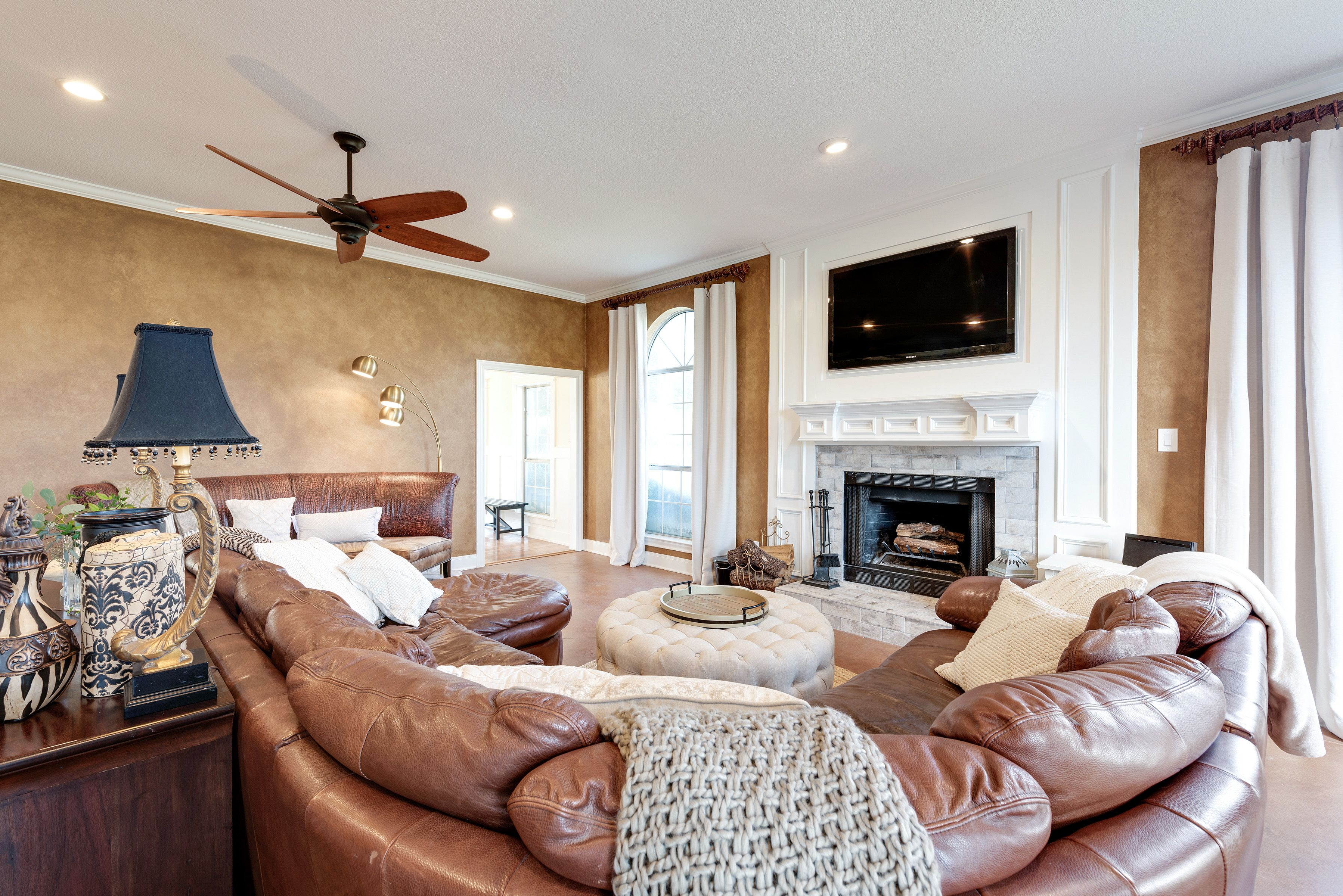 A spacious living room with a leather sofa, a fireplace, a ceiling fan, and a large TV.
