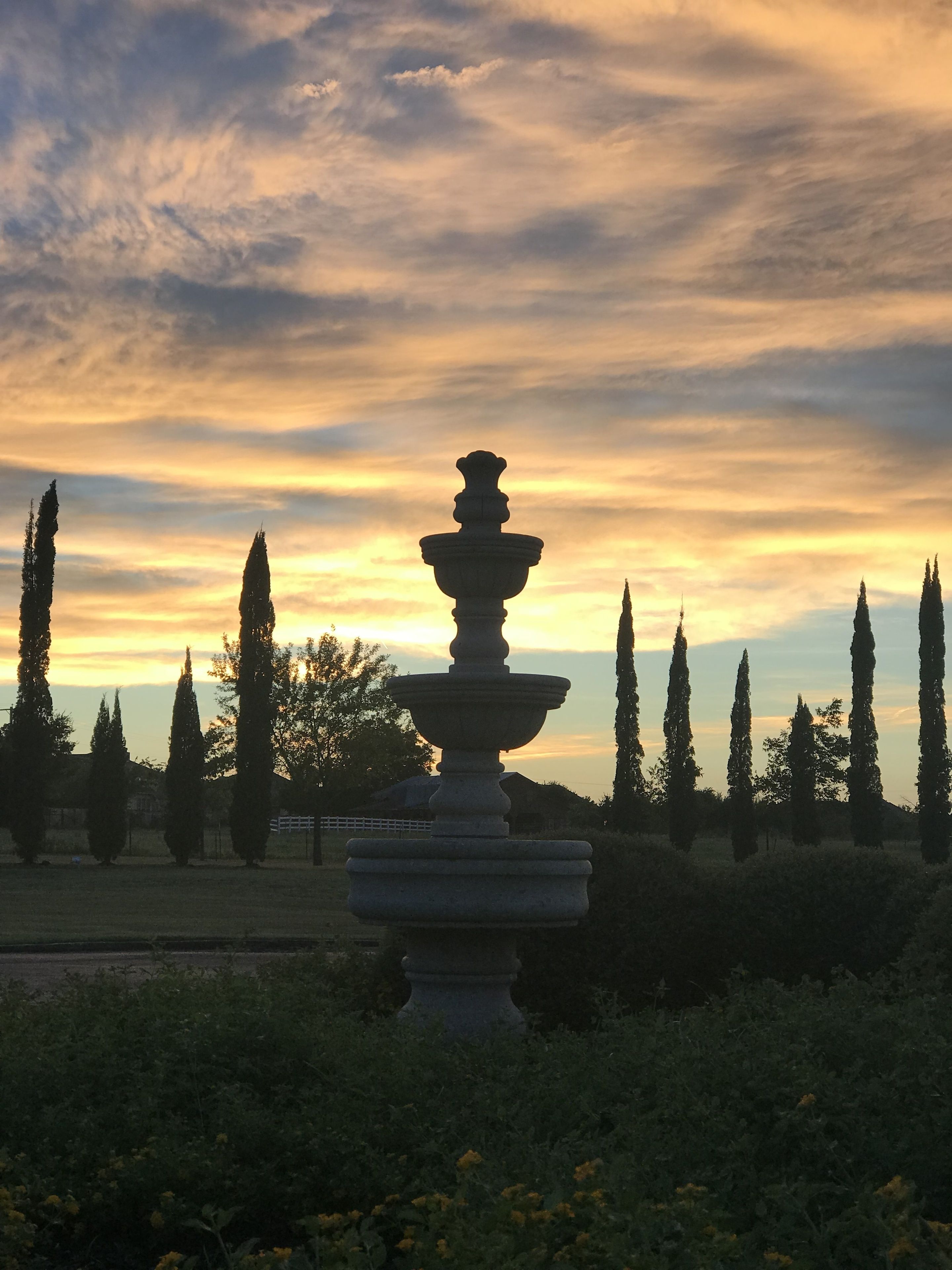 A garden with a central stone fountain, surrounded by tall cypress trees and a sunset sky.