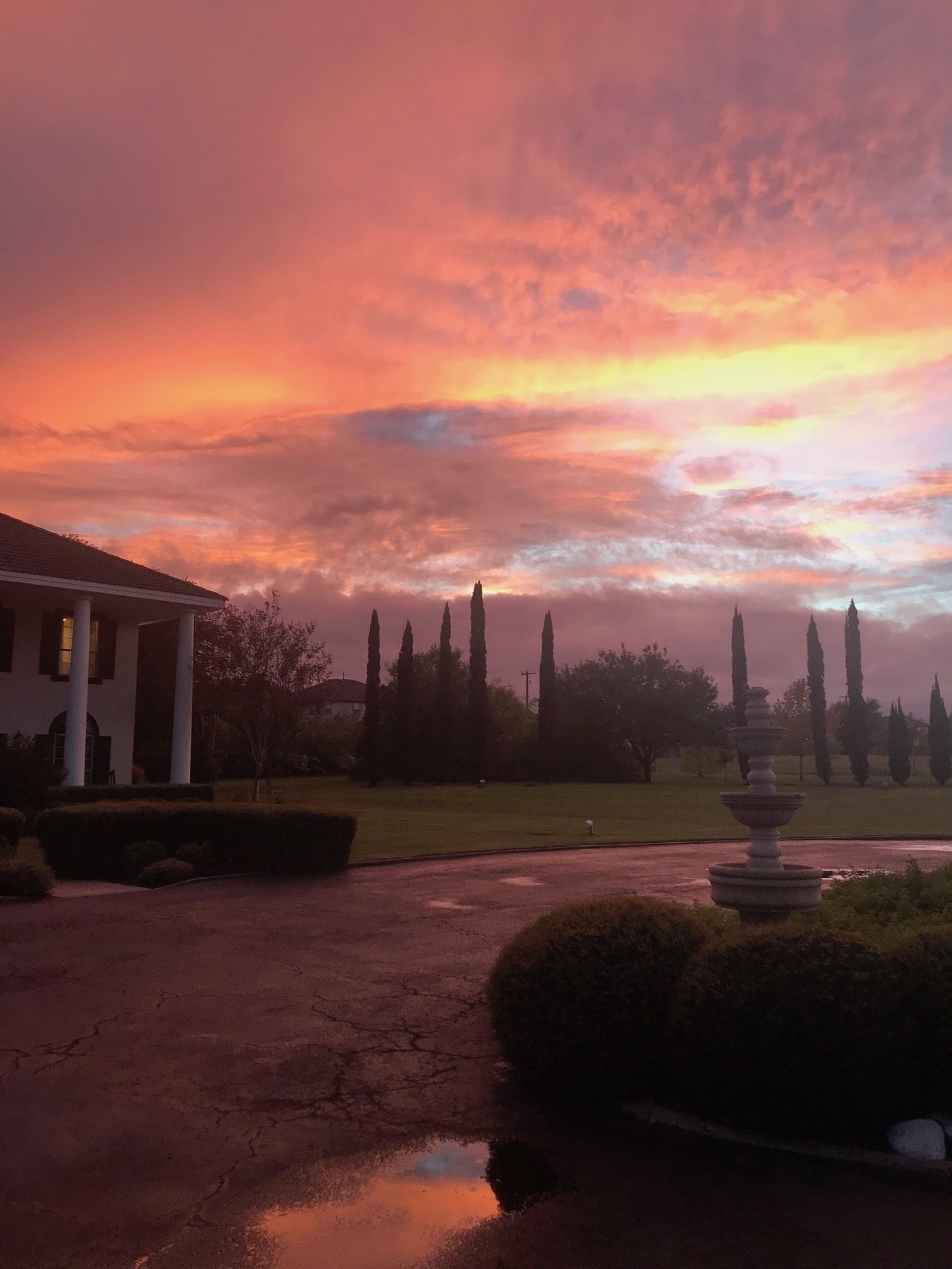 A house with a driveway leading to a fountain, surrounded by trees and a sunset sky.