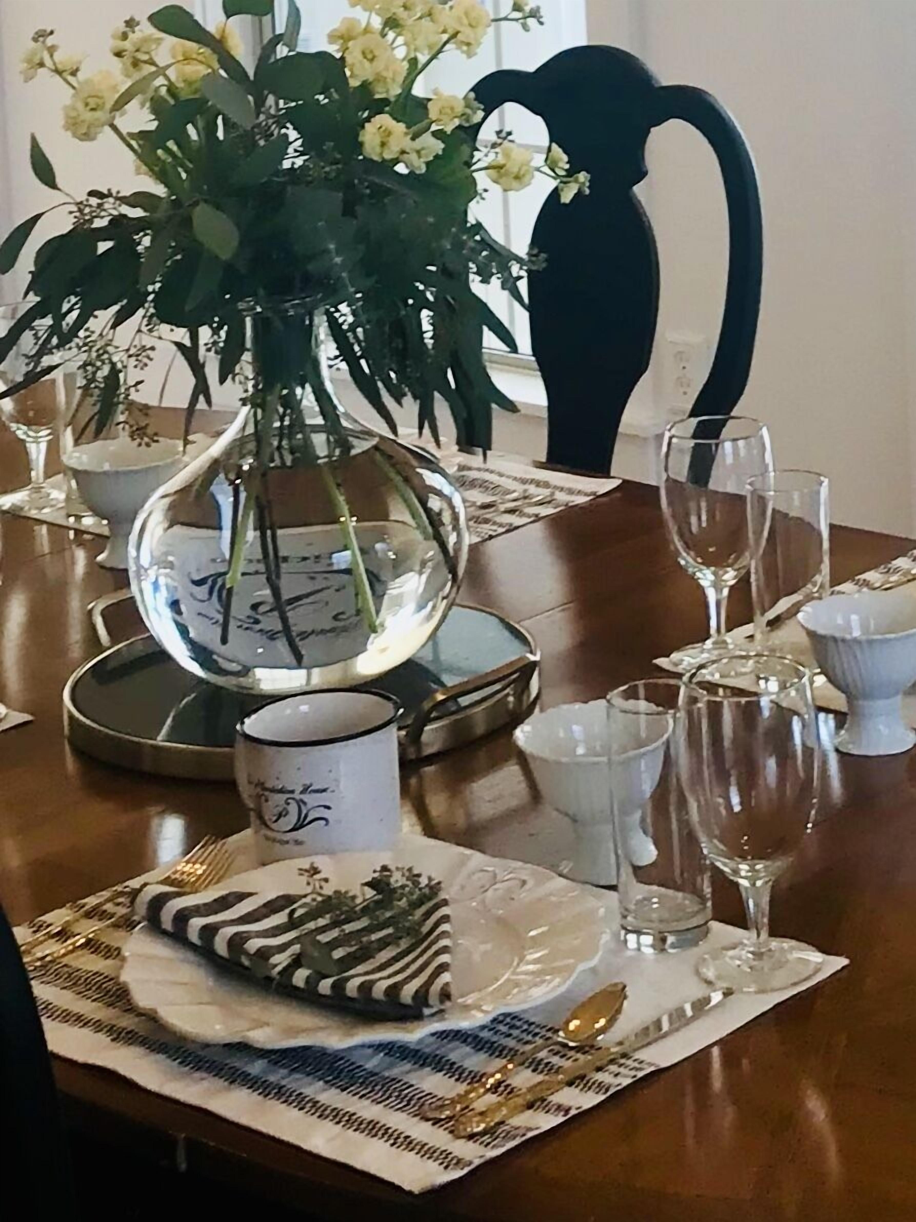 Uma mesa de jantar posta com pratos, talheres e copos, um vaso com flores e um espelho decorativo.
