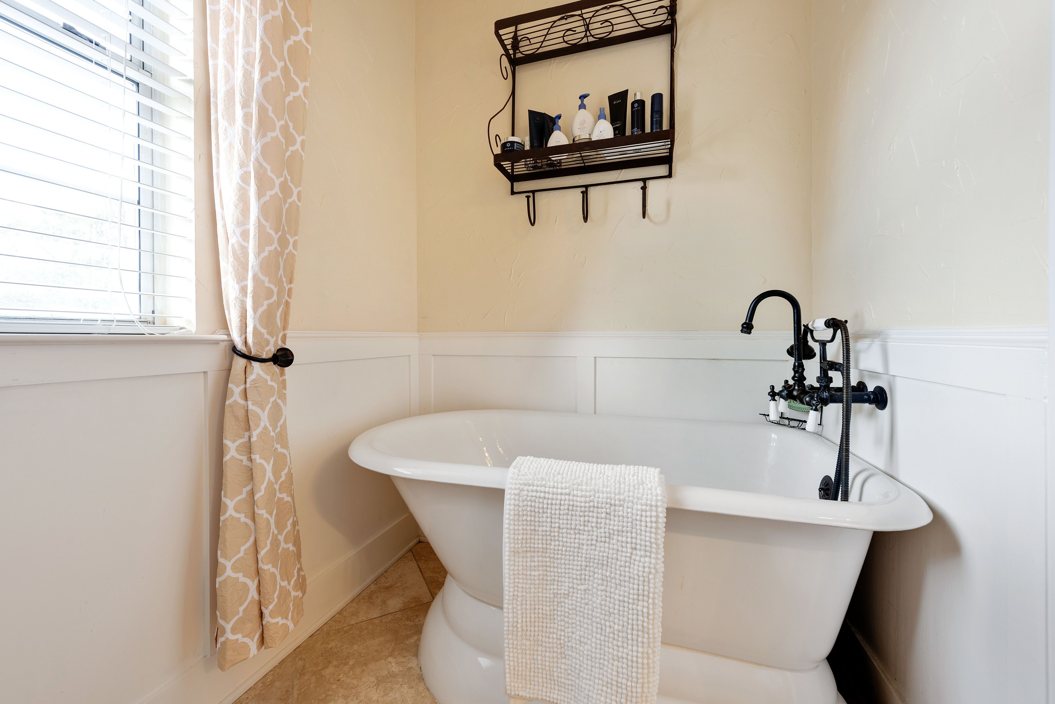 A bathroom with a freestanding bathtub, a towel rack with toiletries, and a window with blinds.