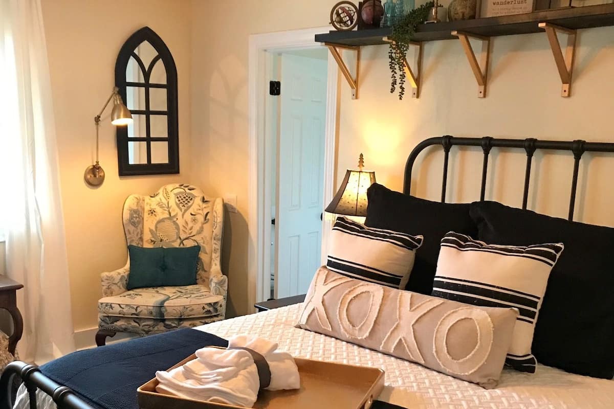 A neatly arranged bedroom with a bed, a chair, a bedside table, and a shelf with decorative items.