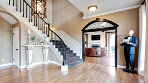 A spacious foyer with a grand staircase, a well-lit living area, and a butler holding a tray.