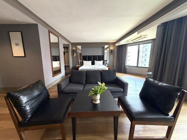 Habitación de hotel con una cama grande, un sofá, dos sillones, una mesita con una planta y un amplio ventanal con cortinas.
