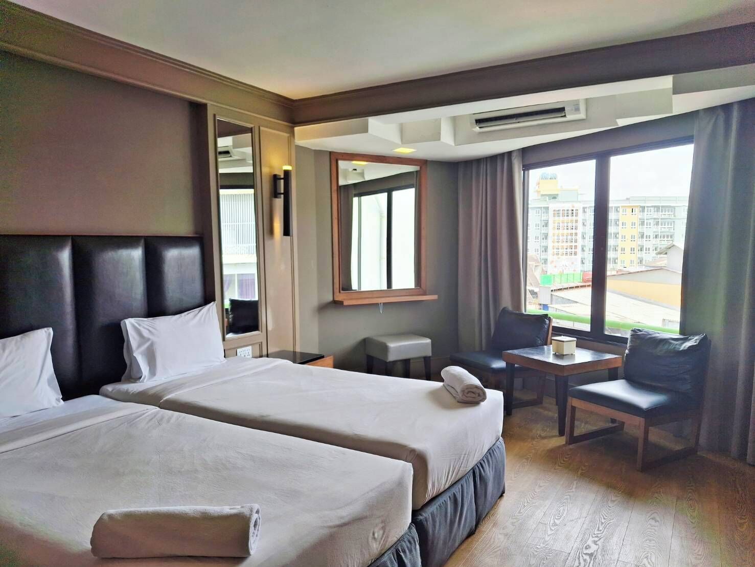 A hotel room with two beds, a small table, a chair, and a large window offering a view of buildings outside.