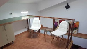 Une petite pièce bien éclairée, avec un plancher en bois, deux chaises blanches, une table en verre et un tableau accroché au mur.