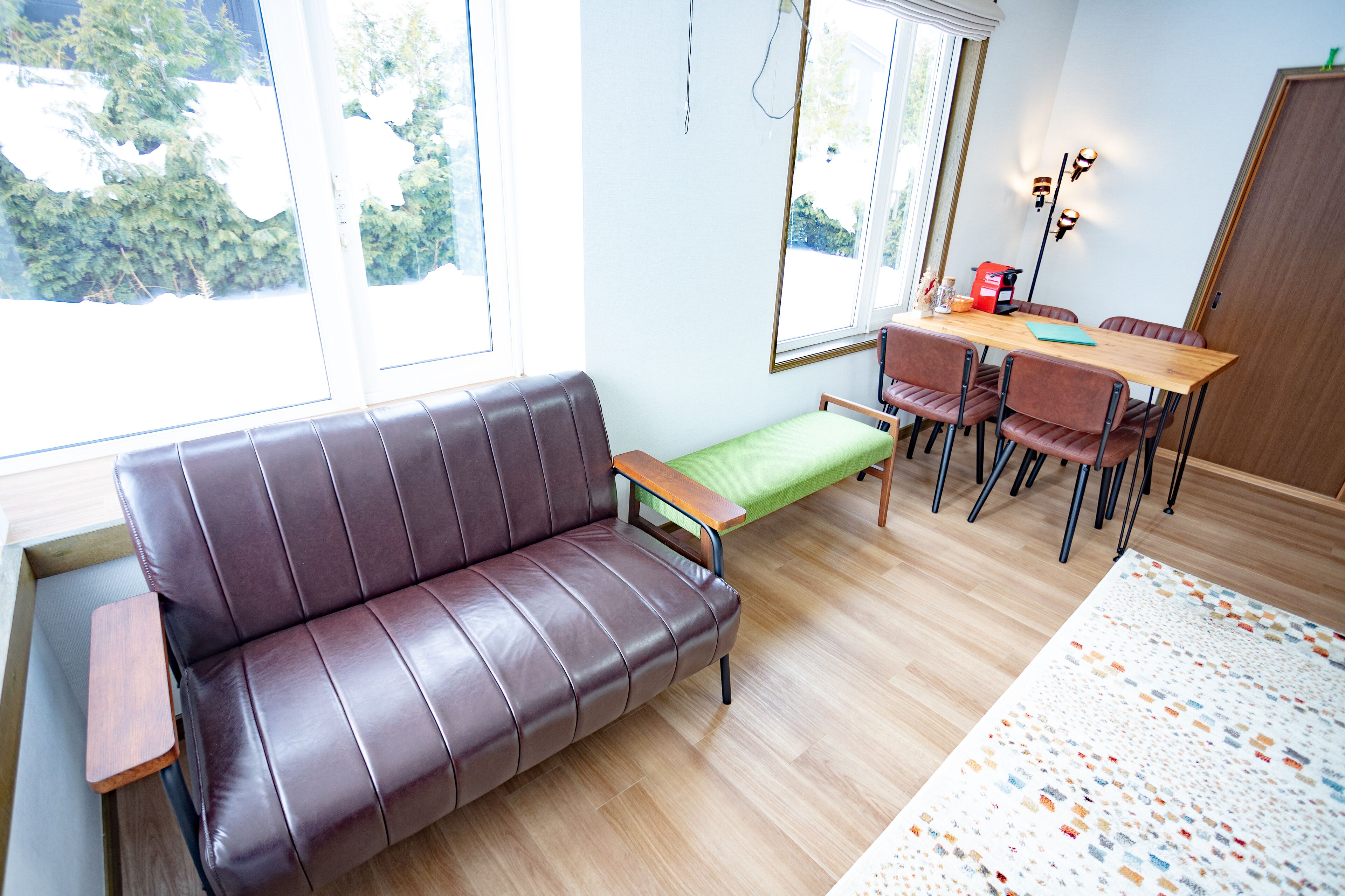 Uma sala de estar moderna com um sofá de couro marrom, uma mesa de centro de madeira, um banco verde e uma área de jantar com mesa e cadeiras de madeira.