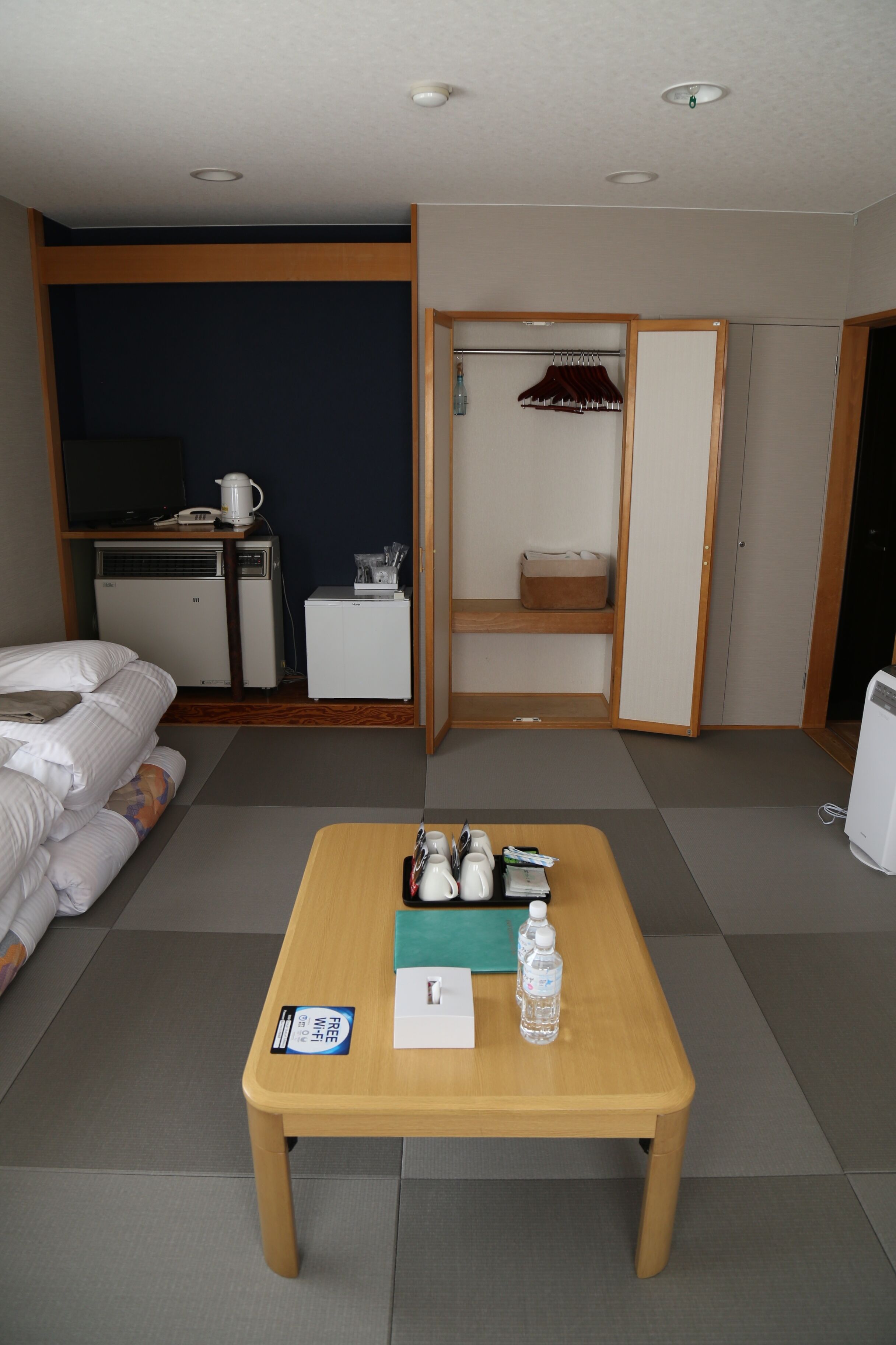 Habitación de hotel con cama, escritorio con ordenador, pequeño refrigerador, mesa de madera con botella de agua y estante con una prenda roja.