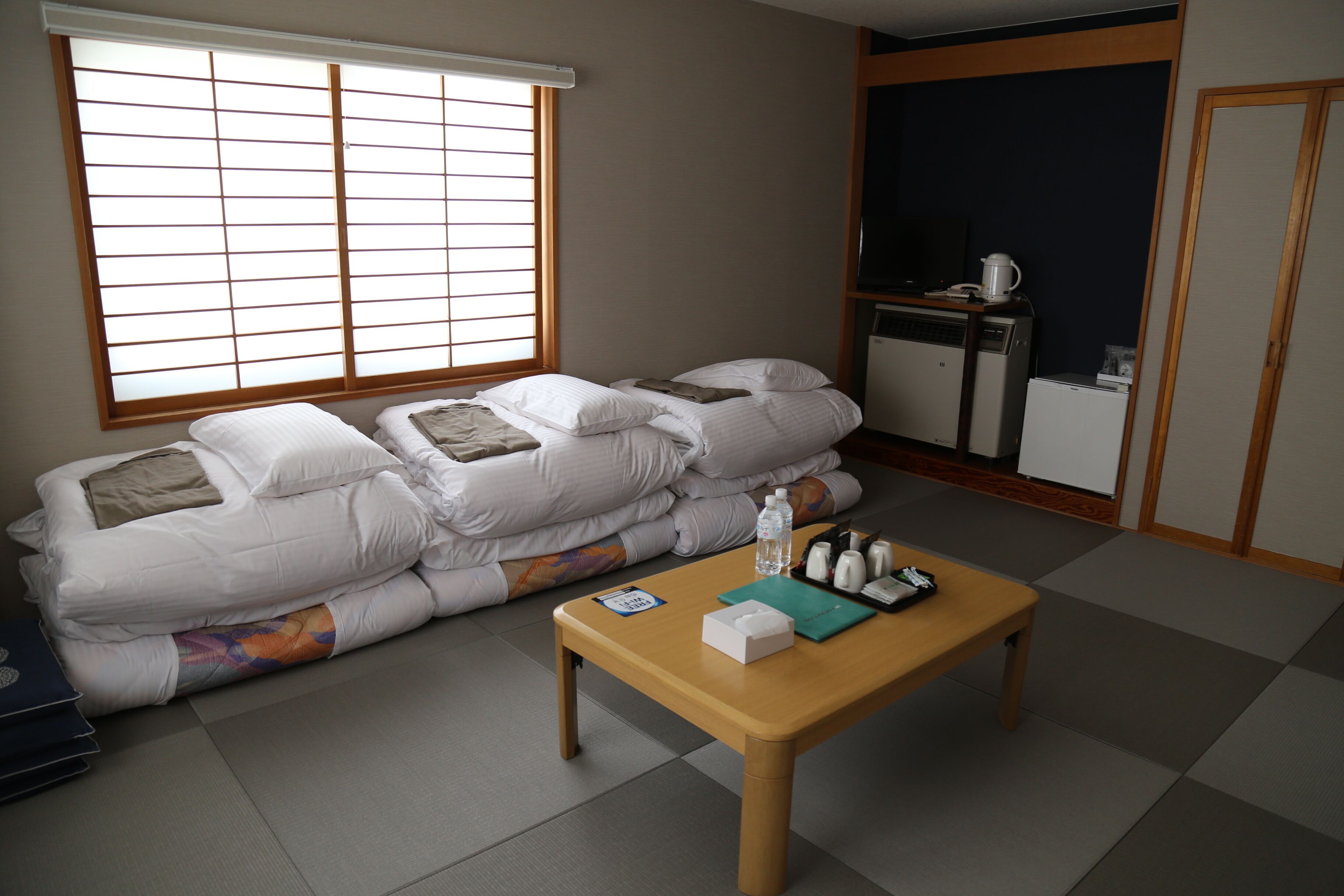Une pièce avec un futon servant de lit, une table en bois, une bouteille d’eau, une télécommande et un petit réfrigérateur.