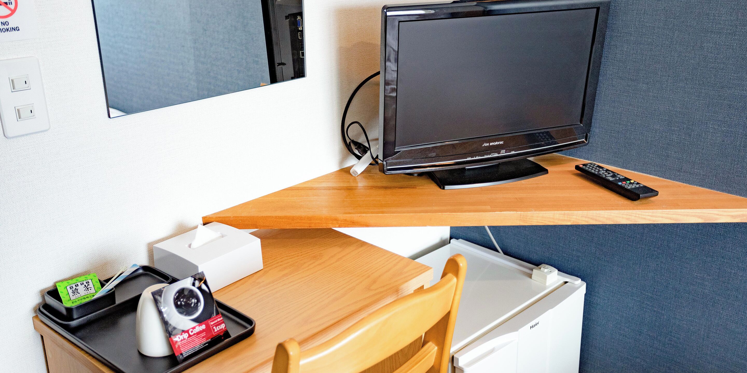 A hotel room with a TV on a wooden desk, a remote control, a camera, and a tray with a coffee cup and a box.