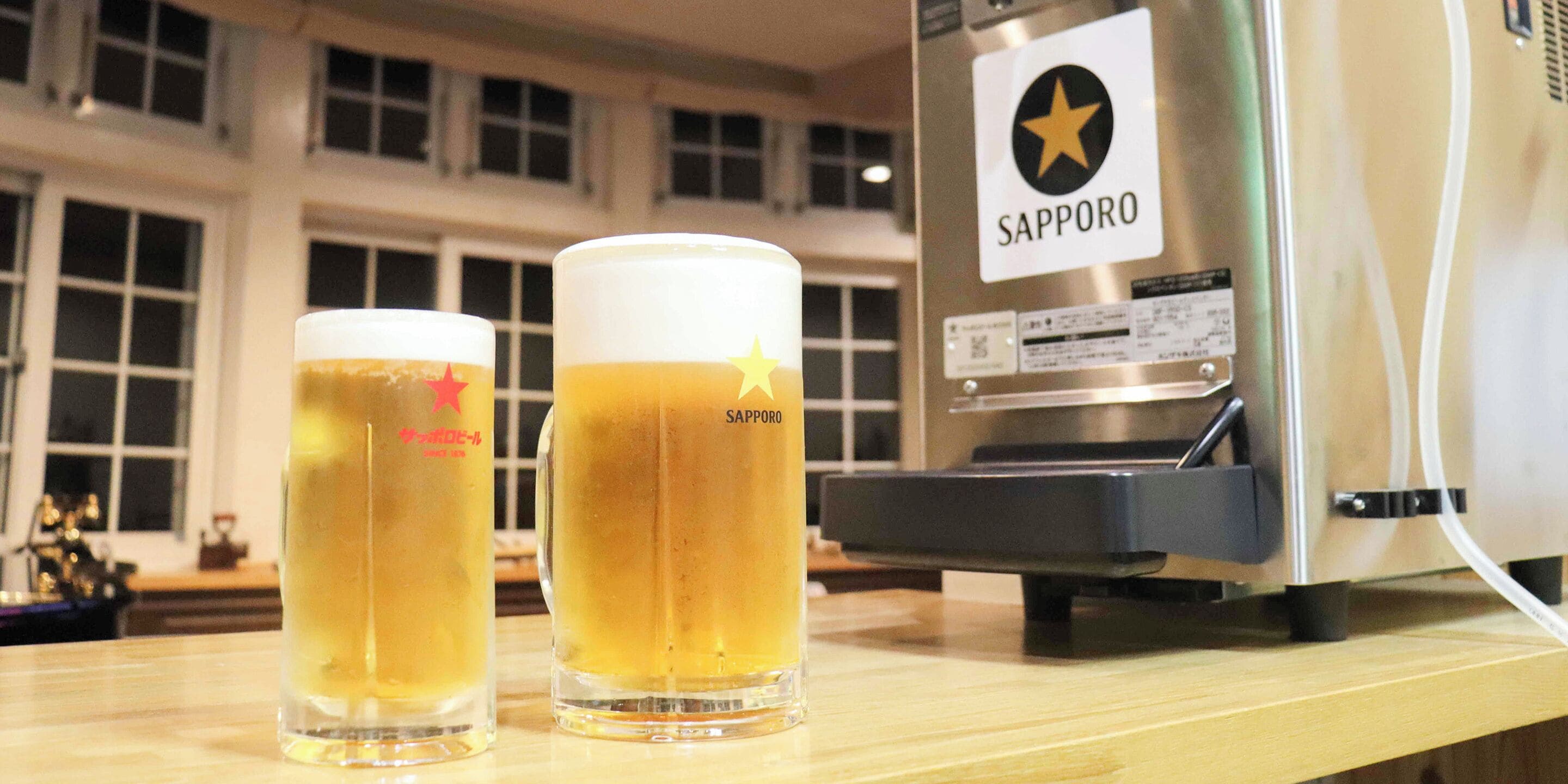 Dois copos de cerveja sobre um balcão de madeira, com uma máquina da marca Sapporo ao fundo.
