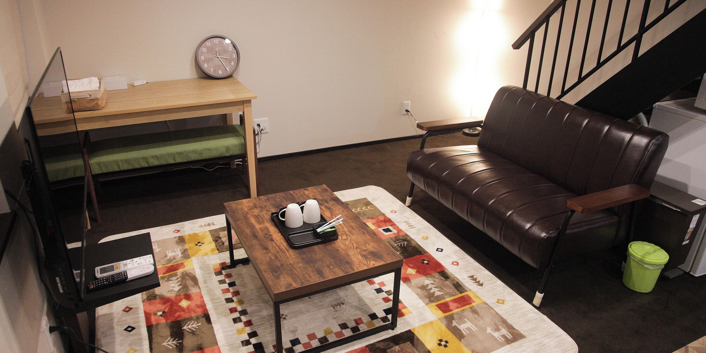 A living room with a brown leather couch, a wooden coffee table with mugs and chopsticks, a clock on the wall, and a TV on a stand.